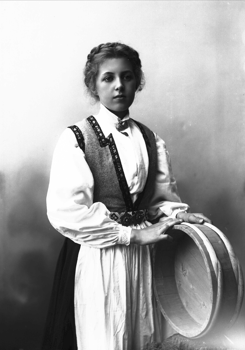 Solveig Lund, 3/4 portrett av kvinne i drakt, tatt i fotografens studio. Hun støtter begge armene mot en melkebunke til høyre i fotografiet. Hun ser ned mot høyre i bildet. Nøytral bakgrunn.
