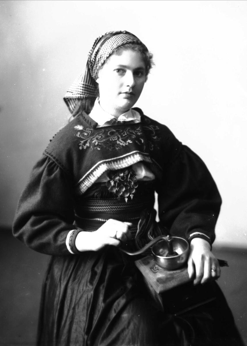 Solveig Lund, 1/2 portrett av kvinne i drakt og skaut, tatt i fotografens studio. Hun sitter og holder en kaffekvern på fanget. Nøytral bakgrunn.