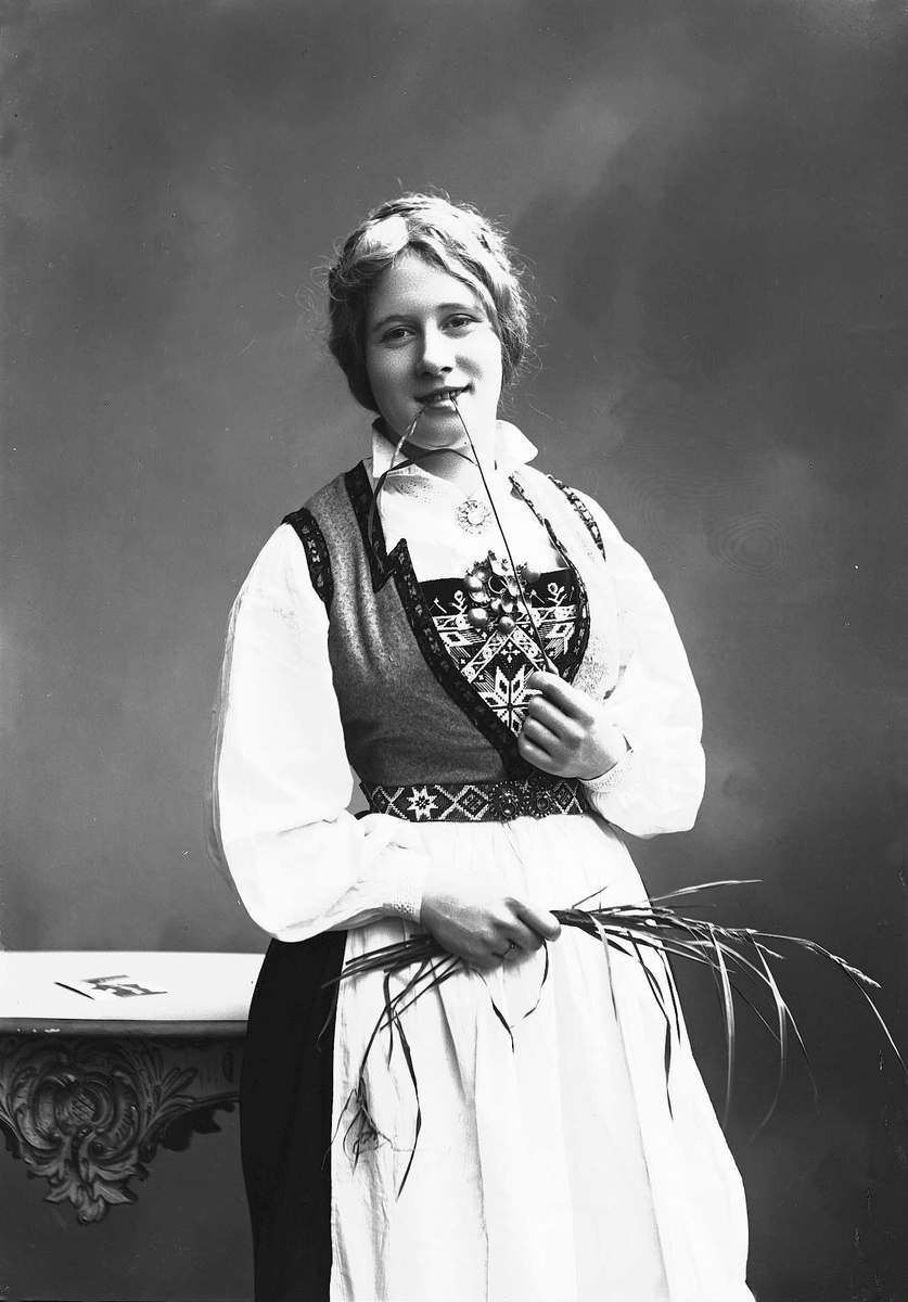 Solveig Lund, 3/4 dels studioportrett av kvinne i drakt og oppsatt hår, som lener seg mot et bord. På bordet ligger det et fotografi. I den høyre hånden holder kvinnen en bunt med strå, i den venstre holder hun et strå som hun tygger på. Nøytral bakgrunn.