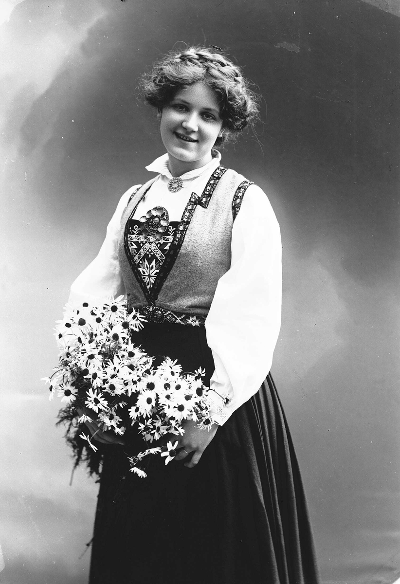 Solveig Lund, 3/4 dels studioportrett av kvinne i drakt og oppsatt hår. I hendene holder hun en favn med prestekrager. Bakgrunnen er malt med himmel og skyer.