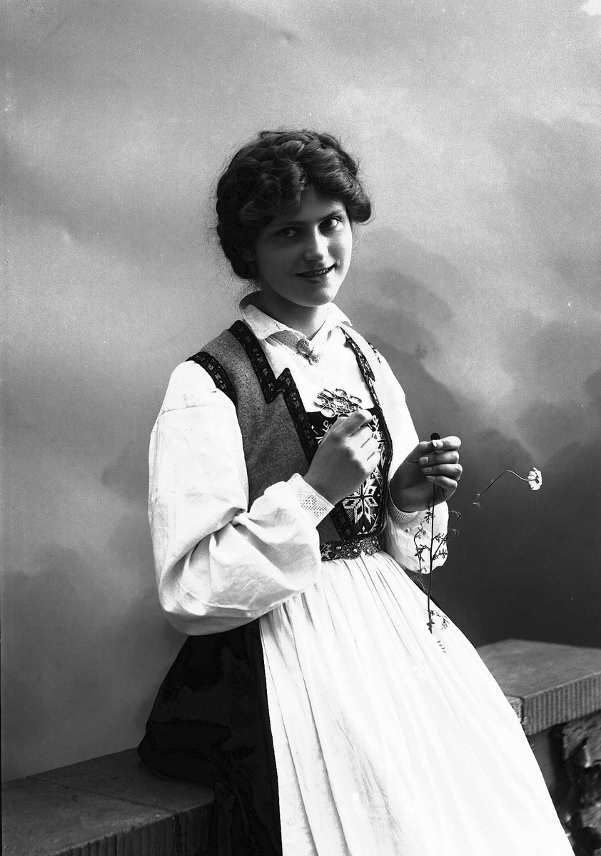Solveig Lund, 3/4 dels studioportrett av kvinne i drakt og oppsatt hår, som sitter på et gjerde. I høyre hånd holder hun en prestekrage. Bakgrunnen er malt med himmel og skyer.