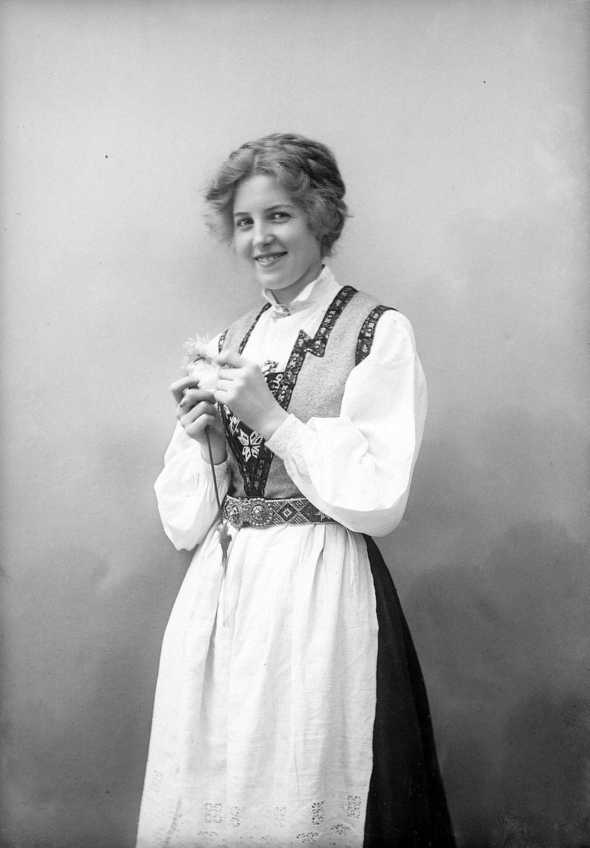 Solveig Lund, 3/4 dels studioportrett av kvinne i drakt og oppsatt hår. I hendene holder hun en blomst. Bakgrunnen er malt med himmel og skyer.
