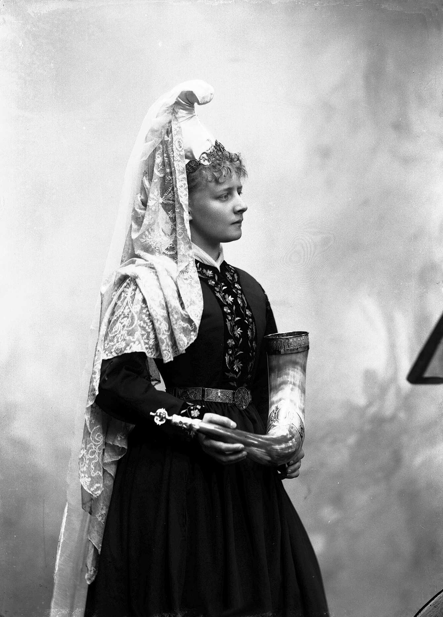 Solveig Lund, 3/4 dels studioportrett av kvinne i drakt og hodeplagg. Hun står foran en nøytral bakgrunn. I hendene holder hun et horn.
