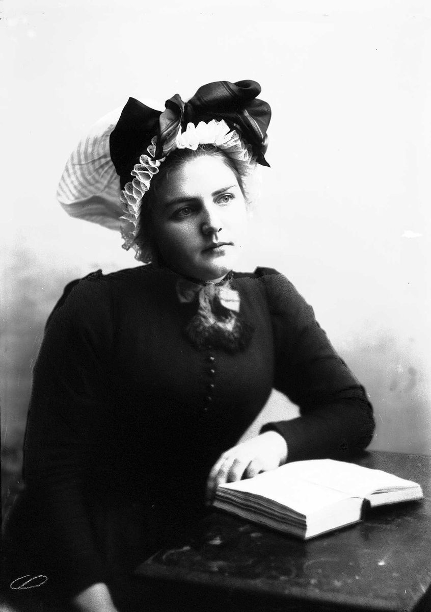 Solveig Lund, 1/2 dels studioportrett av kvinne i drakt, og skaut. hun sitter ved et bord med en oppslått bok. Bakgrunnen er nøytral.