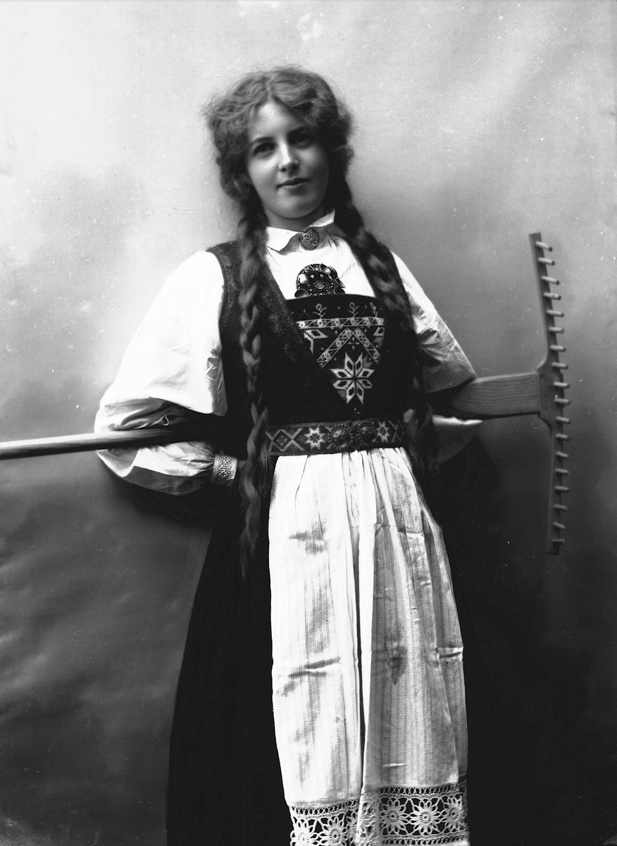 Solveig Lund, 3/4 dels studioportrett av kvinne i drakt, med flettet hår. Hun holder en rive bak ryggen sin. Bakgrunnen er nøytral.