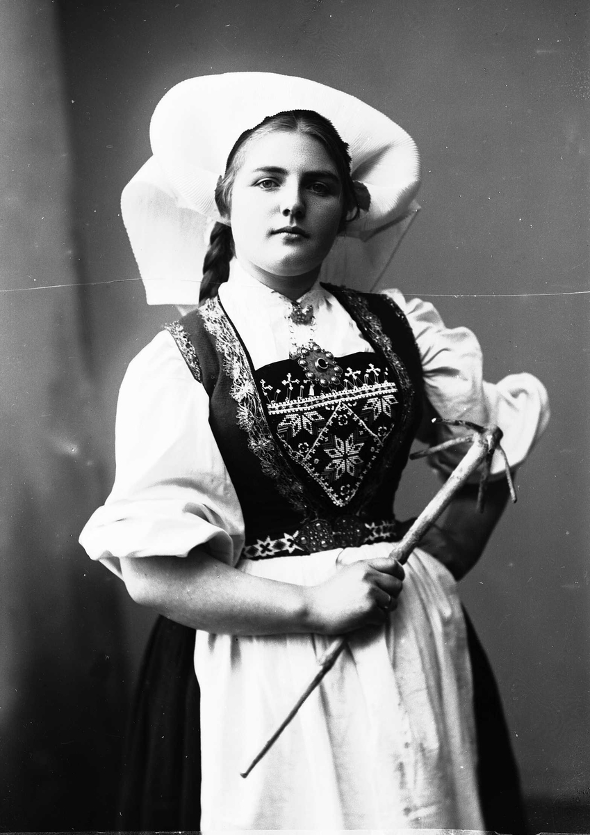 Solveig Lund, 3/4 dels studioportrett av kvinne i drakt, med skaut. Hun holder et redskap i høyre hånd, mens den venstre hviler mot hoften. Bakgrunnen er nøytral.