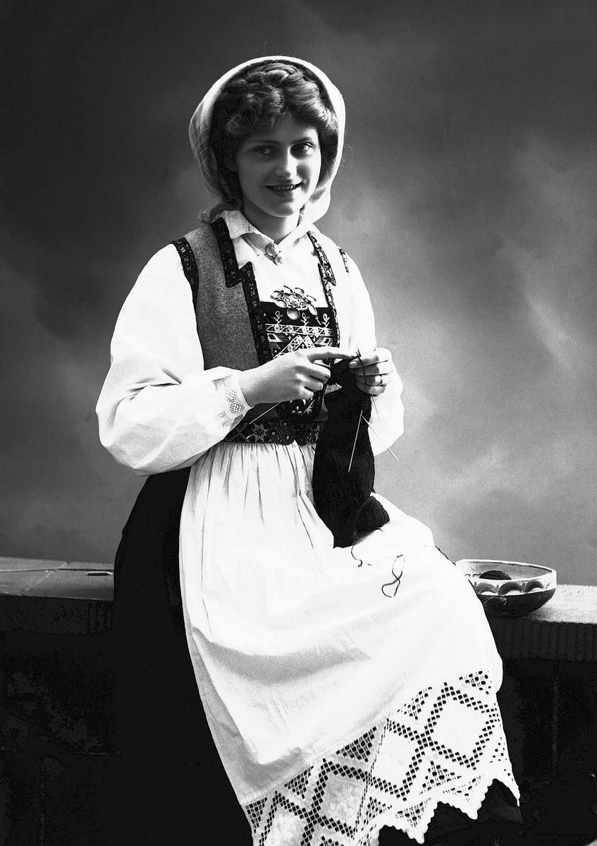Solveig Lund, 3/4 dels studioportrett av kvinne i drakt og skaut, som sitter på en benk og strikker. Til høyre for henne står en bolle. Bakgrunnen er nøytral.