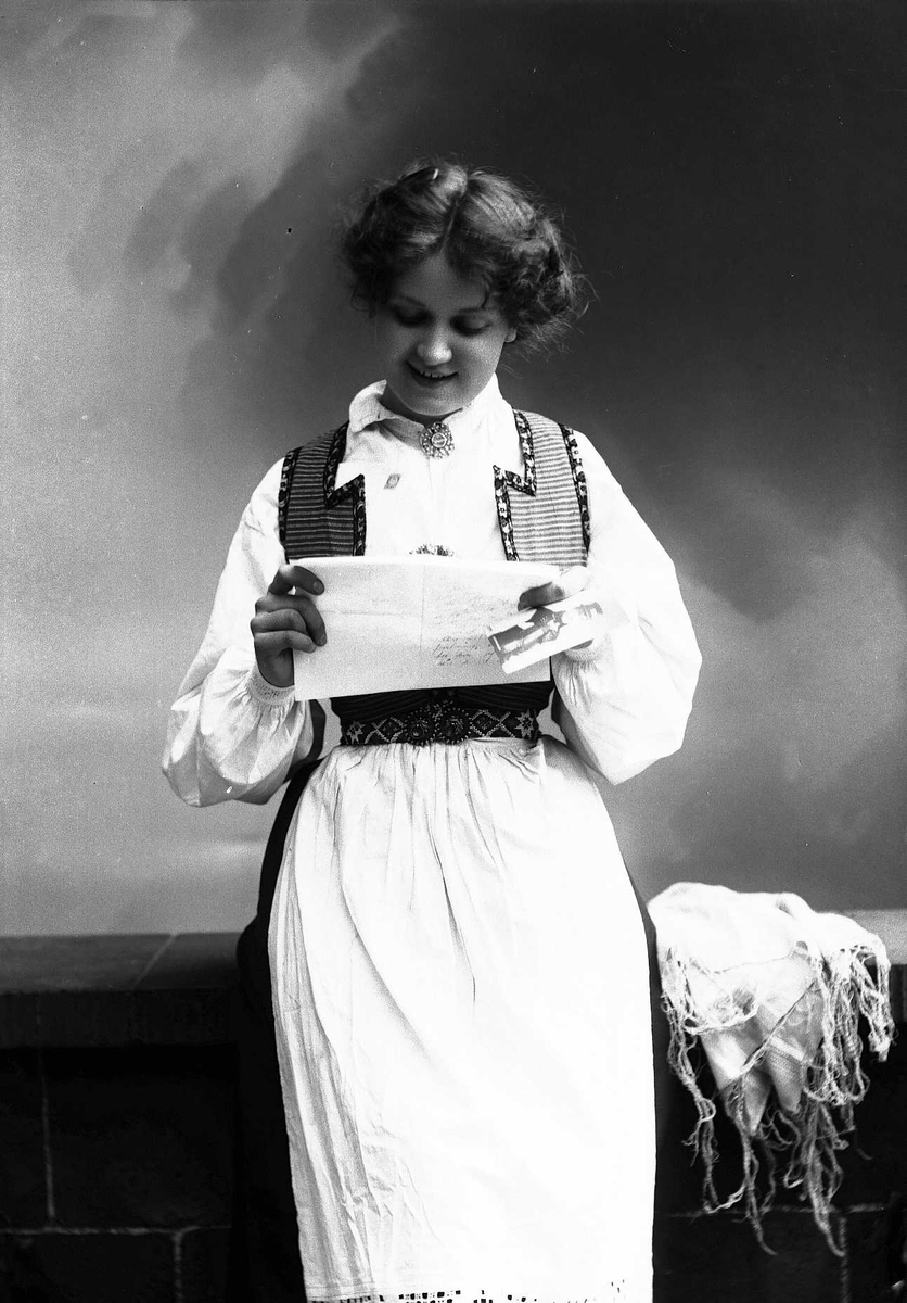 Solveig Lund, 3/4 dels studioportrett av kvinne i drakt, som sitter på en benk og leser et brev. I venstre hånd holder hun et fotografi av en mann. På benken til venstre for henne ligger et sjal. Bakgrunnen er nøytral.