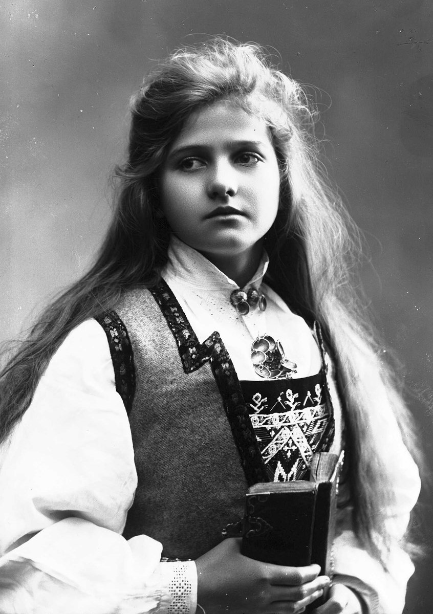 Solveig Lund, 1/2 dels studioportrett av kvinne i drakt med utslått hår. Hun holder en bok i hendene. Bakgrunnen er nøytral.
