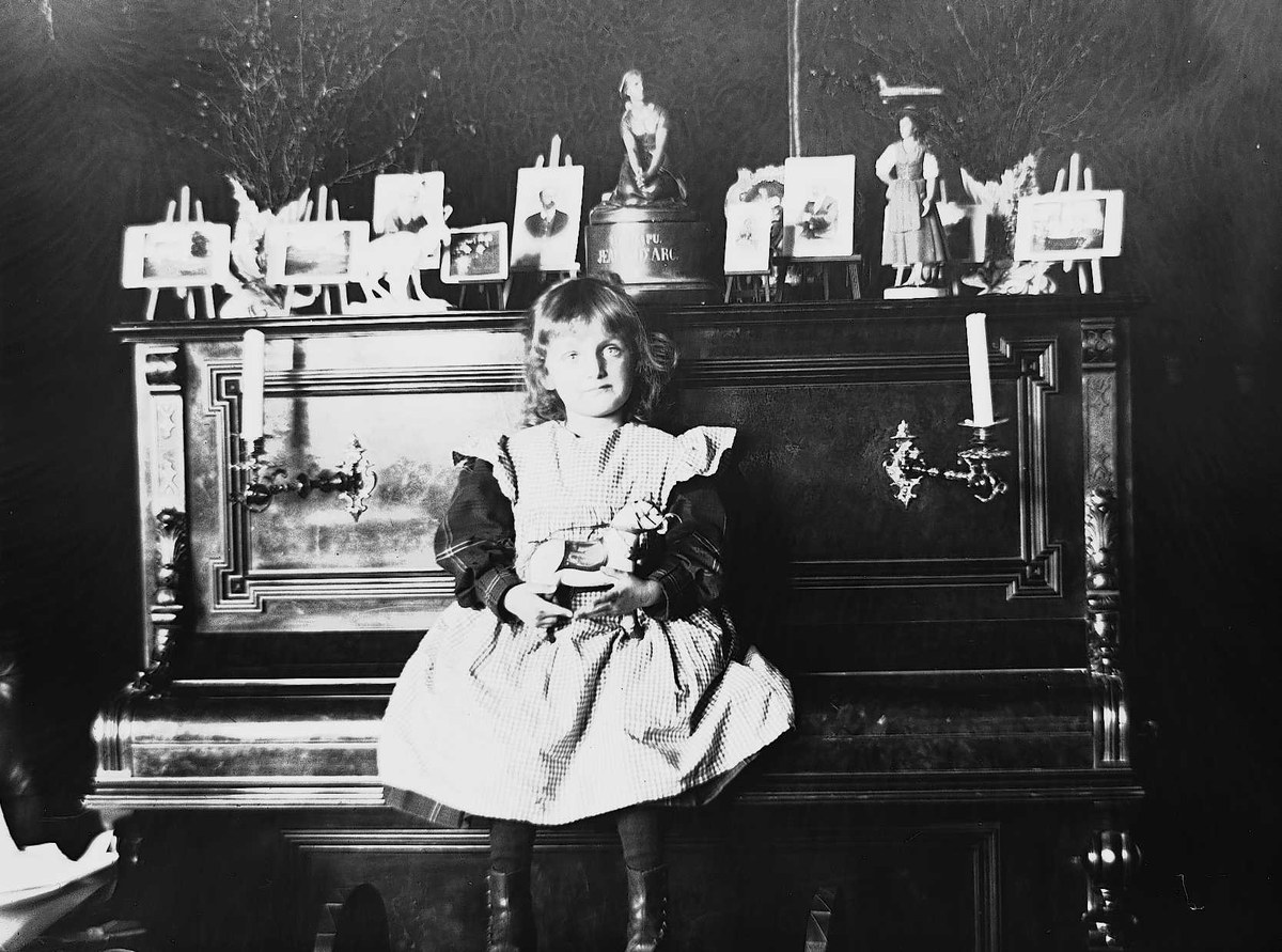 Axel Quinsgaard Wiborg, Karen Q. Wiborg sitter på piano i stue, Meltzers gate 9, Oslo, 1899. Fotografier i ramme plassert på pianoet.