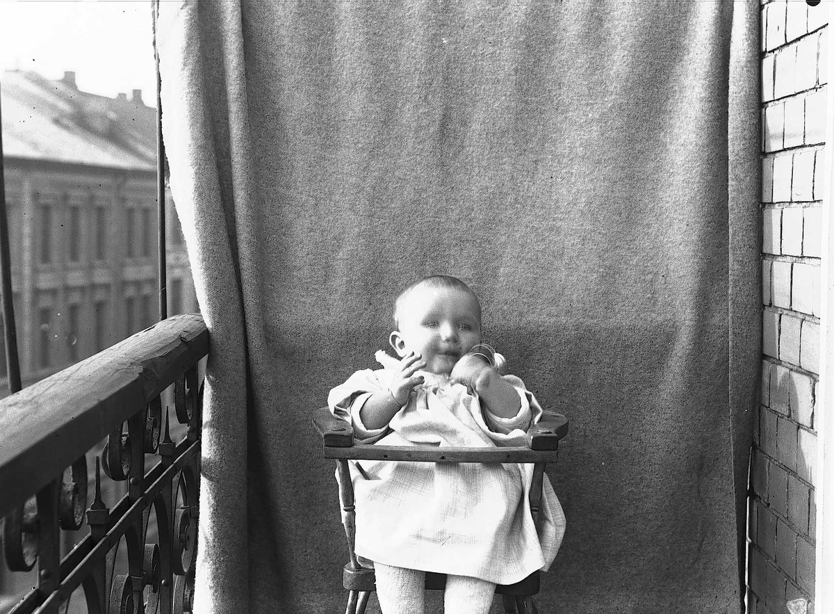 Axel Quinsgaard Wiborg, Lillebror Axel Q. Wiborg i barnestol på balkong, ant. Meltzers gate 9, Oslo, 1899. Fotografert mot opphengt bakteppe.