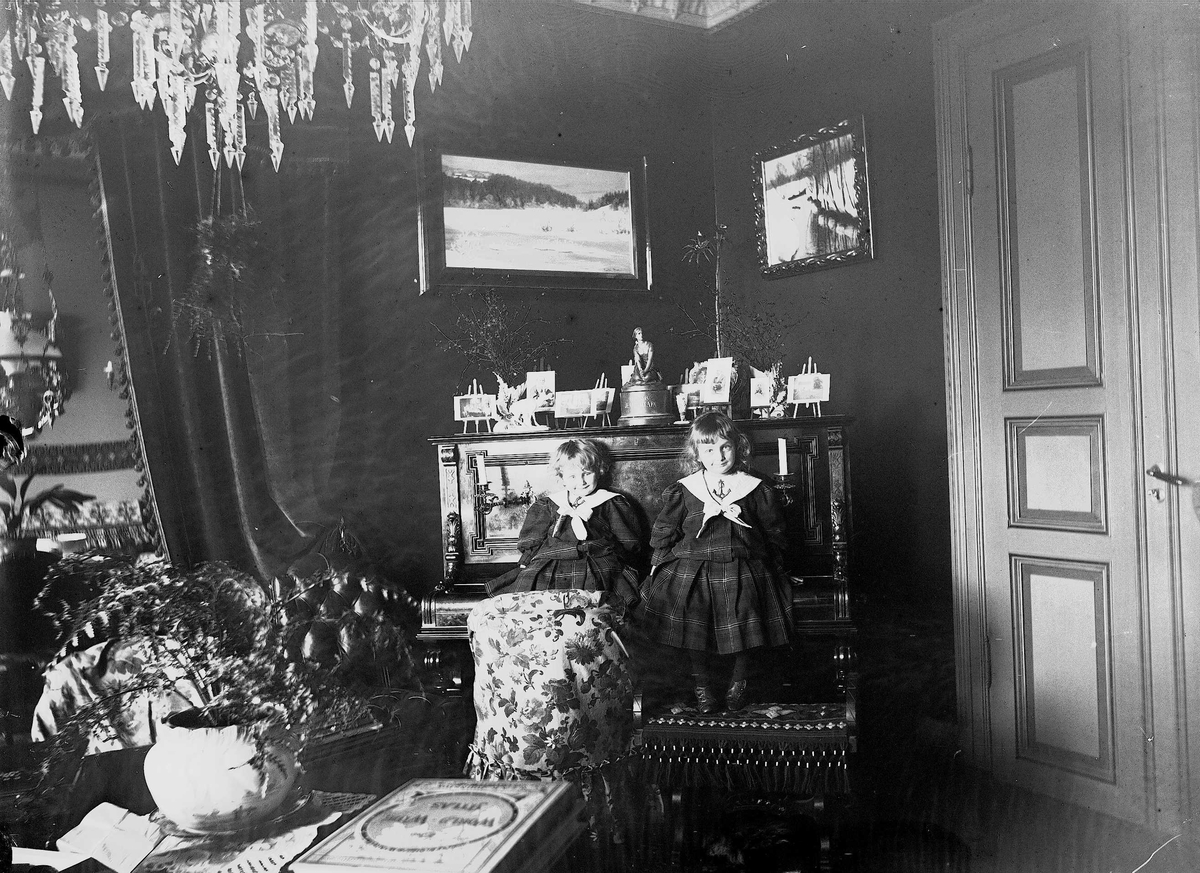 Axel Quinsgaard Wiborg, Gudrun og Karen Q. Wiborg sitter på piano i stue, Meltzers gate 9, Oslo, 1899. Fotografier i ramme plassert på pianoet.
