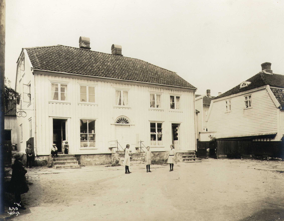 Anders Beer Wilse, Gammelt trehus med butikker i første etasje, Lillesand, Aust-Agder. Barn leker på plassen foran.