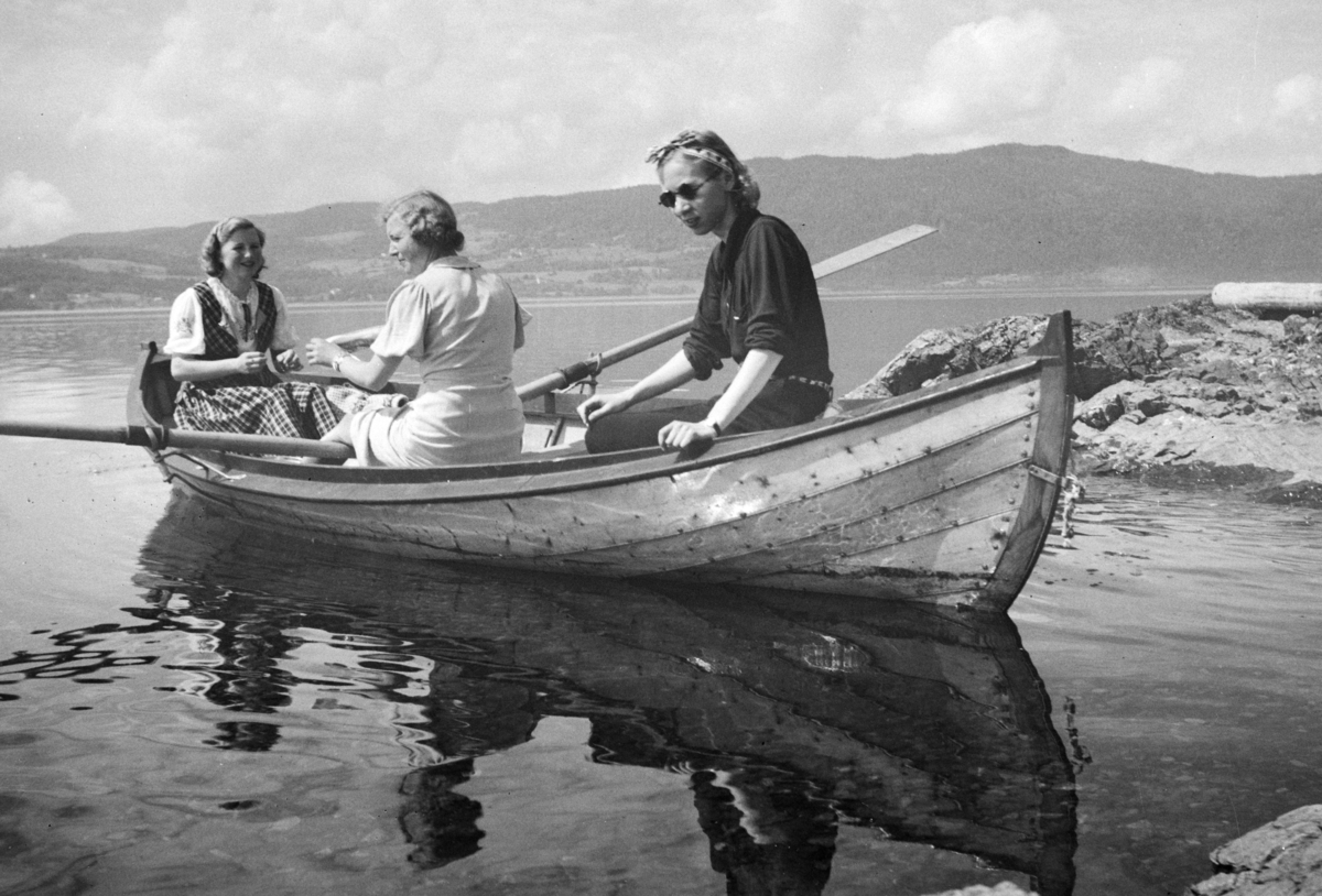 Johs. (Johannes) Johannessen, Robåt (dreggebåt, mjøsbåt, Hollandbåt), Til høyre sitter Lumi Johannessen to ukjente, utenfor "Lia, " hytta til Baker Johs. Johannessen på Jessnes i Furnes. Mjøsa. Båten er ikke malt(ant. lakkert), har utliggere for årene, typiske for mange av mjøsbåtene.