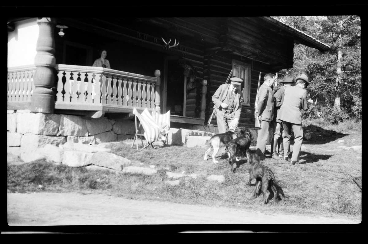 Rolf Sr. Sundt, Jegere med hunder utenfor jakthytte. Rolf Sundt Sr. med gevær og hunder, Johan Buratti samt to ukjente. Ukjent kvinne på verandaen. Fotografert 1928.