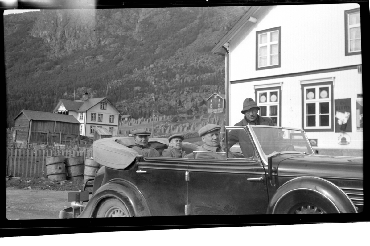 Rolf Sr. Sundt, Johan Buratti, Erling Lorck sitter bak i bilen (Buick 1934-35 convertible sedan) mens Harsem sitter i forsetet sammen med stående Rolf Sundt sr., Vannevik, Hemsedal. To gårder ligger i bakgrunnen . Fotografert 1938.