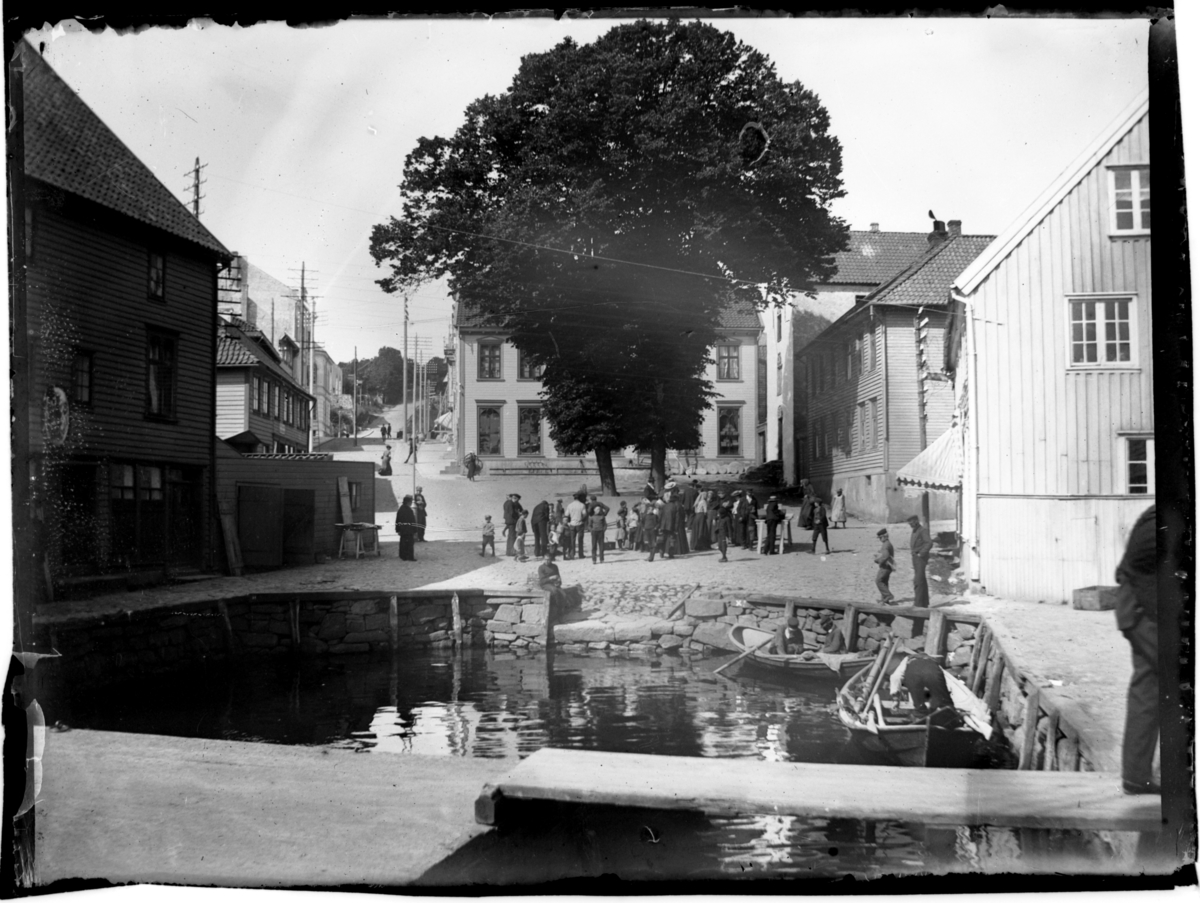 Rolf Sr. Sundt, Farsund torv med småbåthavnen i forgrunnen. En gruppe mennesker er samlet på torvet. Fotografert 1903.