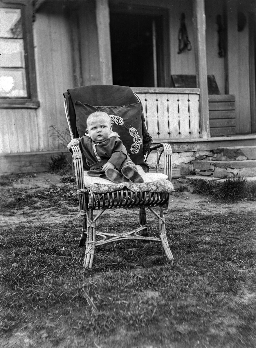 Ole M Martinsen, En baby sitter i en stol utenfor inngangen til et hus. Barnet er Erling Olsen, født 18.06-1924 i Løten. Bildet er tatt ute på tunet foran huset på Vollestadstuen i Løten. Egils foreldre var Ole og Karen Edvardsen ( Vollestadstuen)