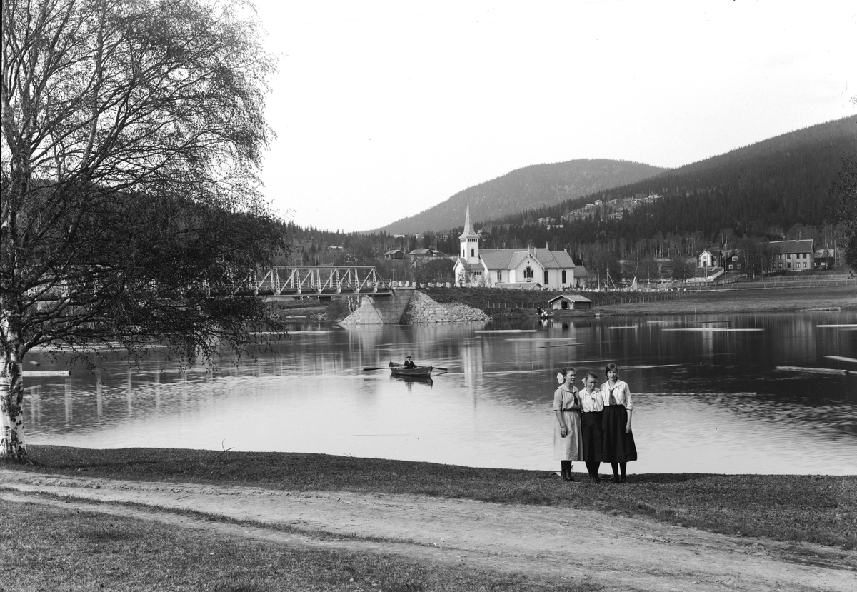 Jakob Jakobsen Engerdal, Innbygda fotografert fra vestsiden av Trysilelva, kvinnene i forgrunnen ukjente. Vi ser Kirkebroen, Trysil hovedkirke og Trysil prestegård. Foran kirken ses taket på atelieret til fotograf G. G. Berg.
