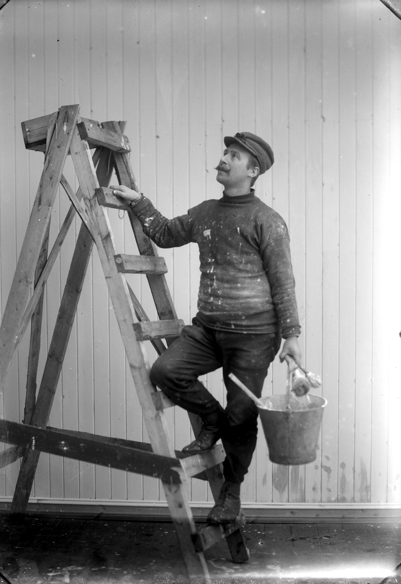 Jakob Jakobsen Engerdal, Maler står i gardintrapp foran panelvegg, med egenblandet maling i sinkbøtte. Malerkoster, klær med malingsflekker.