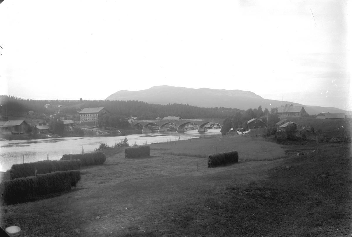 Jakob Jakobsen Engerdal, Nybergsund. Fotografert fra østsida av Trysilelva mot nordvest, Trysilfjellet i bakgrunnen. Brua sto fra 1879-1929. På vestsida av elva ses Nybakk og Nyberg, huset i to etasjer på østsida som ligger delvis skjult bak en høyde er Folkvang.