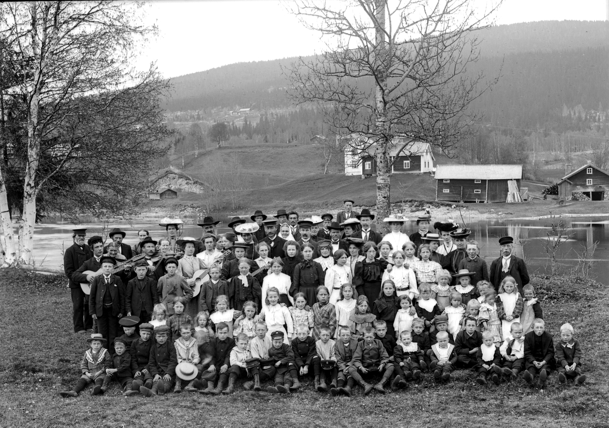 Jakob Jakobsen Engerdal, Stor forsamling, barn, kvinner og menn, foran elva. I bakgrunnen ligger Nordby, Innbygda. Musikkinstrumenter, gitarer (enkel og dobbelgitar/harpegitar), fele.