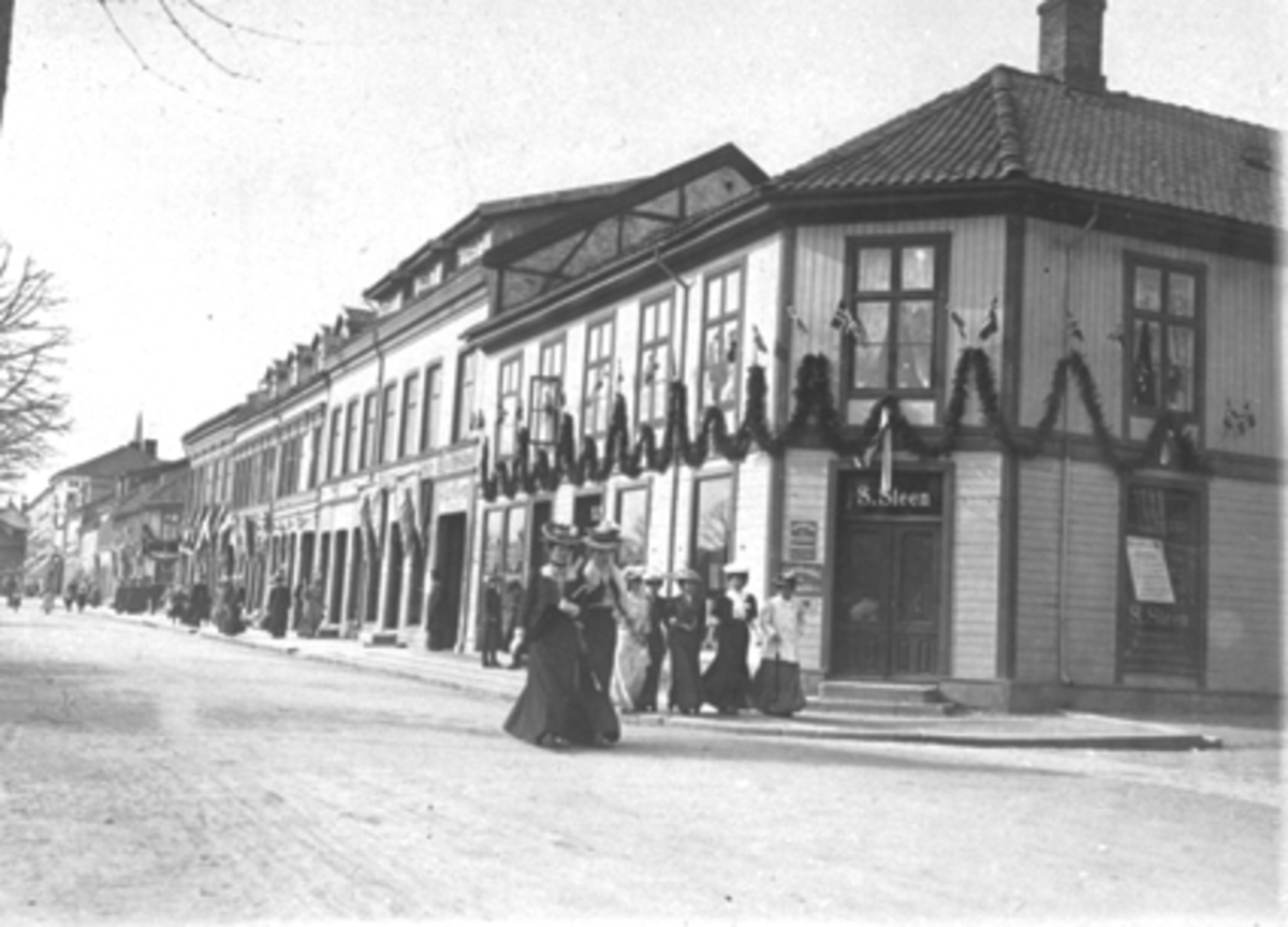 Anno Domkirkeodden, STRANDGATA 61, S. STEENS FORRETNING PYNTET MED GIRLANDERE OG NORSKE FLAGG, KVINNER I KJOLER