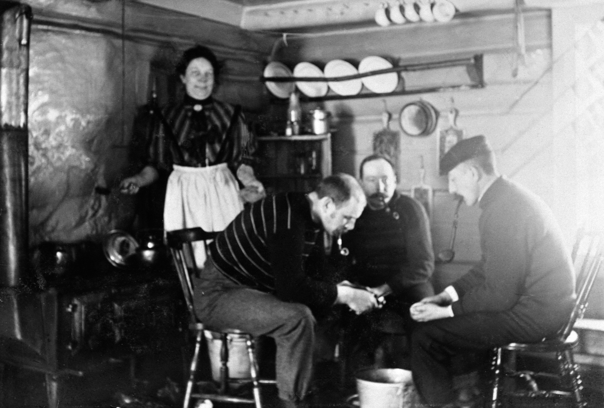 Anno Domkirkeodden, Interiør, kjøkken med vedkomfyr. Tre menn med piper skreller poteter. Dame (kokka) ser på. I midten Philip N. Petersen, Furnesåsen.