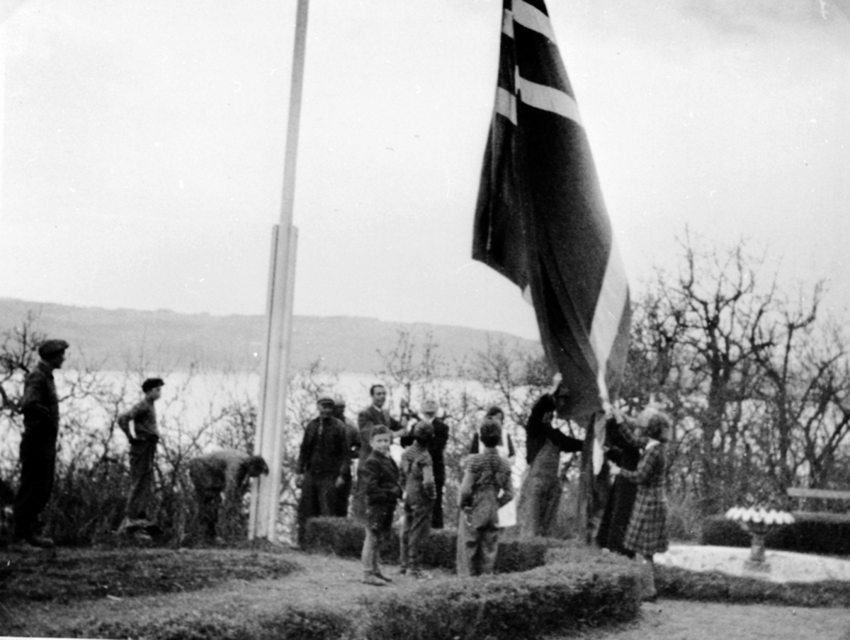 Anno Domkirkeodden, Stor gruppe barn og voksne rundt flaggstange ved flaggheising fredsdagene i mai 1945. Nils Olssøn, eldstesønnen på garden fikk æren av å heise flagget. Prester som var tvangsflyttet deltok. Hovelsrud, Helgøya.