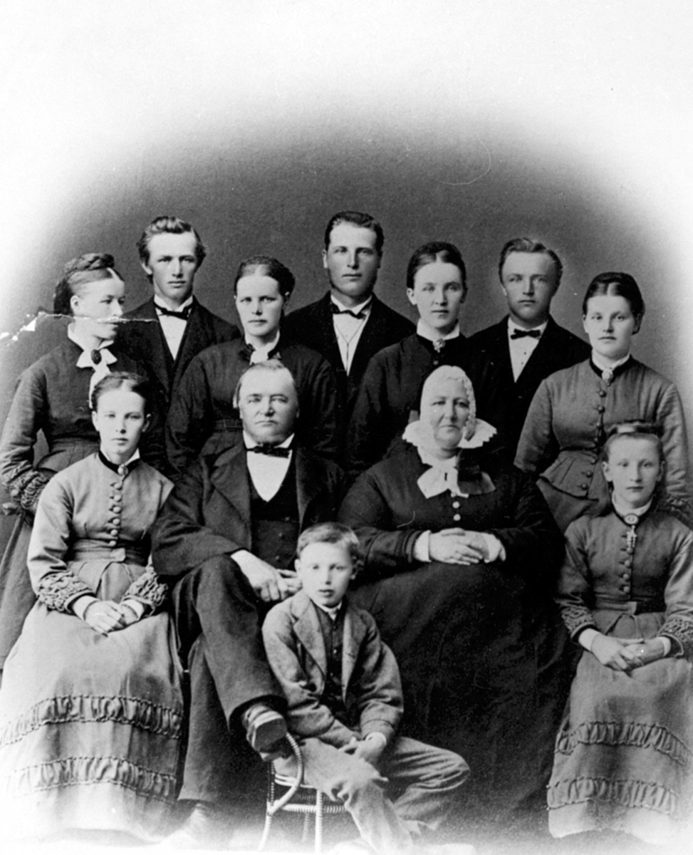 Anno Domkirkeodden, Familiegruppe, Narud gard 1875. Berthe (1823-1903) og Johannes Narud (1812-1904) med 10 av sine 13 barn. Bak fra venstre er Lisbeth, Mathias, Karen, Lars, Maria, Gustav, Louise. Foran fra venstre er Julie, Johannes, Berthe, Anna Birgitte. Forna sitter Arne, Johannes Arnesen Narud var født på Torsæt…