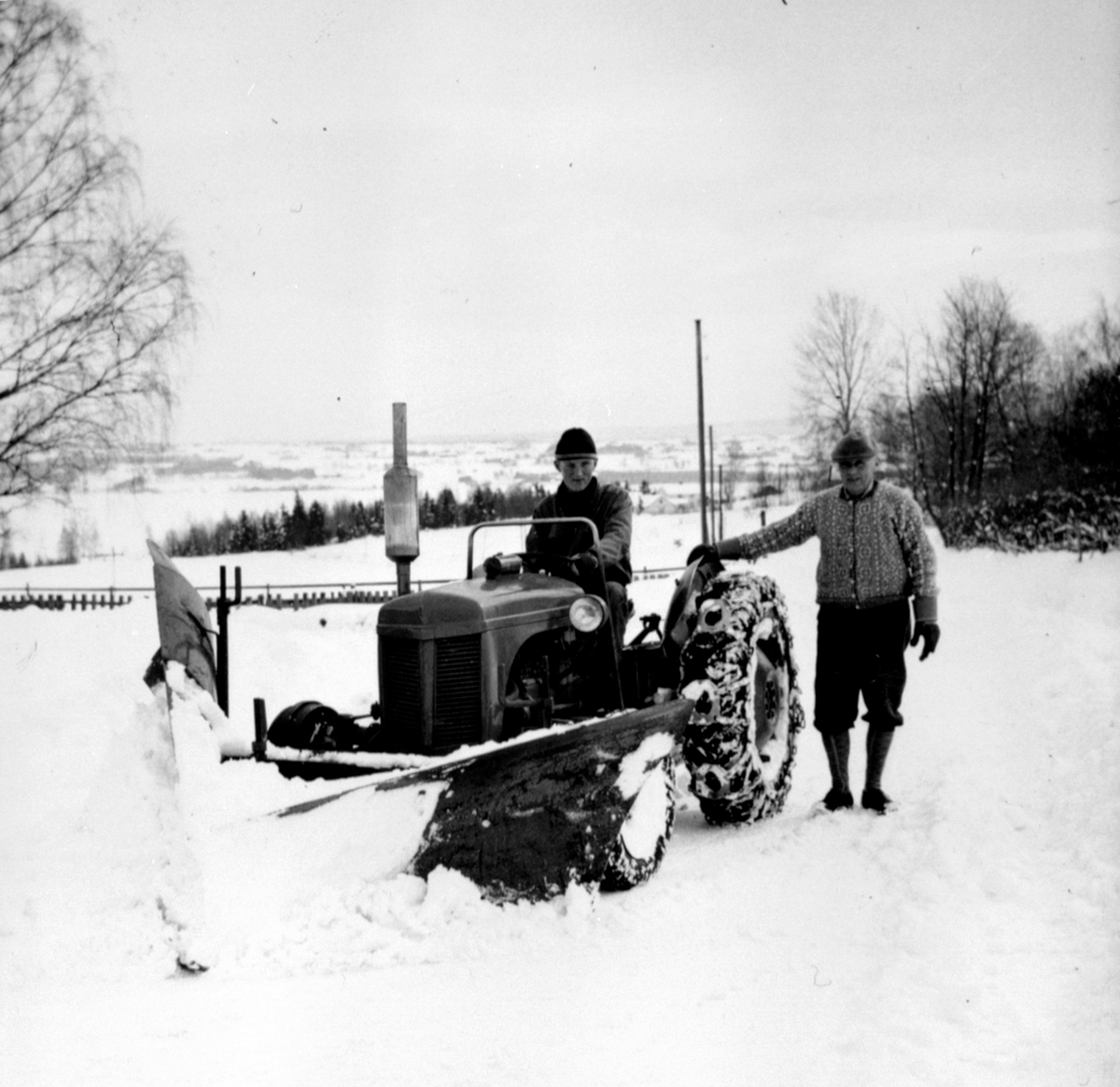 Anno Domkirkeodden, Traktor med snøplog i Lodviken, Helgøya. Bård Dobloug kjører traktoren, Mattis Dobloug står ved siden av. Gråtass.