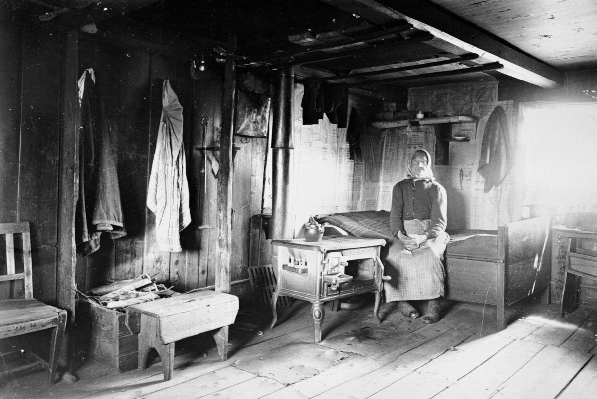 Anno Domkirkeodden, Interiør, husmannsplass, kone på senga, Eline Stueløkken, Stuggulykkja under Bårdseth nedre, Ringsaker. Vedkomfyr ved senga, aviser på veggene som isolasjon. Ca. 1905.