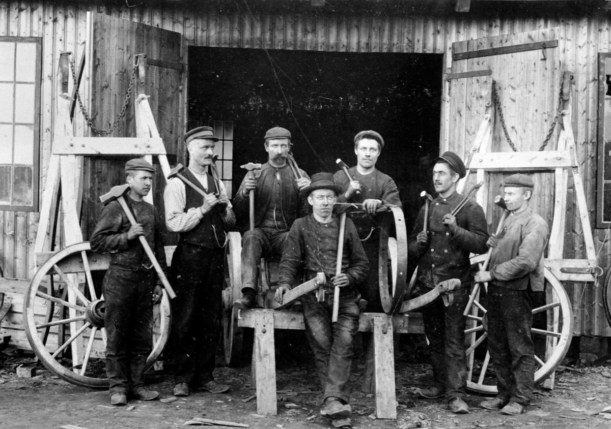 Anno Domkirkeodden, Gruppe smeder utenfor smia på Moelven Brug. Produksjon av kjerrehjul. Fra venstre er Ole Hansen, Lars Pedersen, Tolleif Skau smedmester, Gustav Eliassen, Karl Knutsen, Kristian Skar, Johannes Bekkeslåtten. Karl Knutsen var fra Løten.