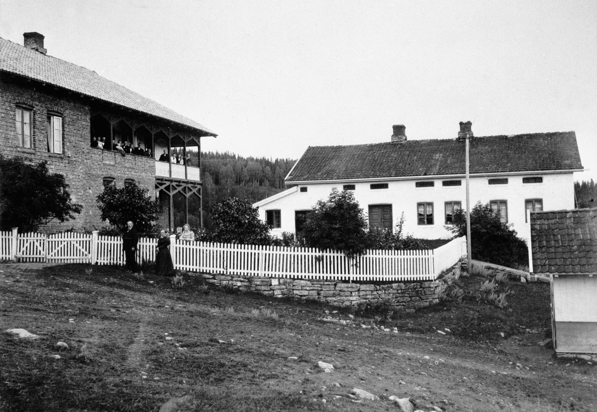 Anno Domkirkeodden, Hovedbygning og føderådsbygning på Mengshoel, Nes, Hedmark. Foran stakittgjerdet er Lise og Jens Mengshoel. Mye folk på veranda.