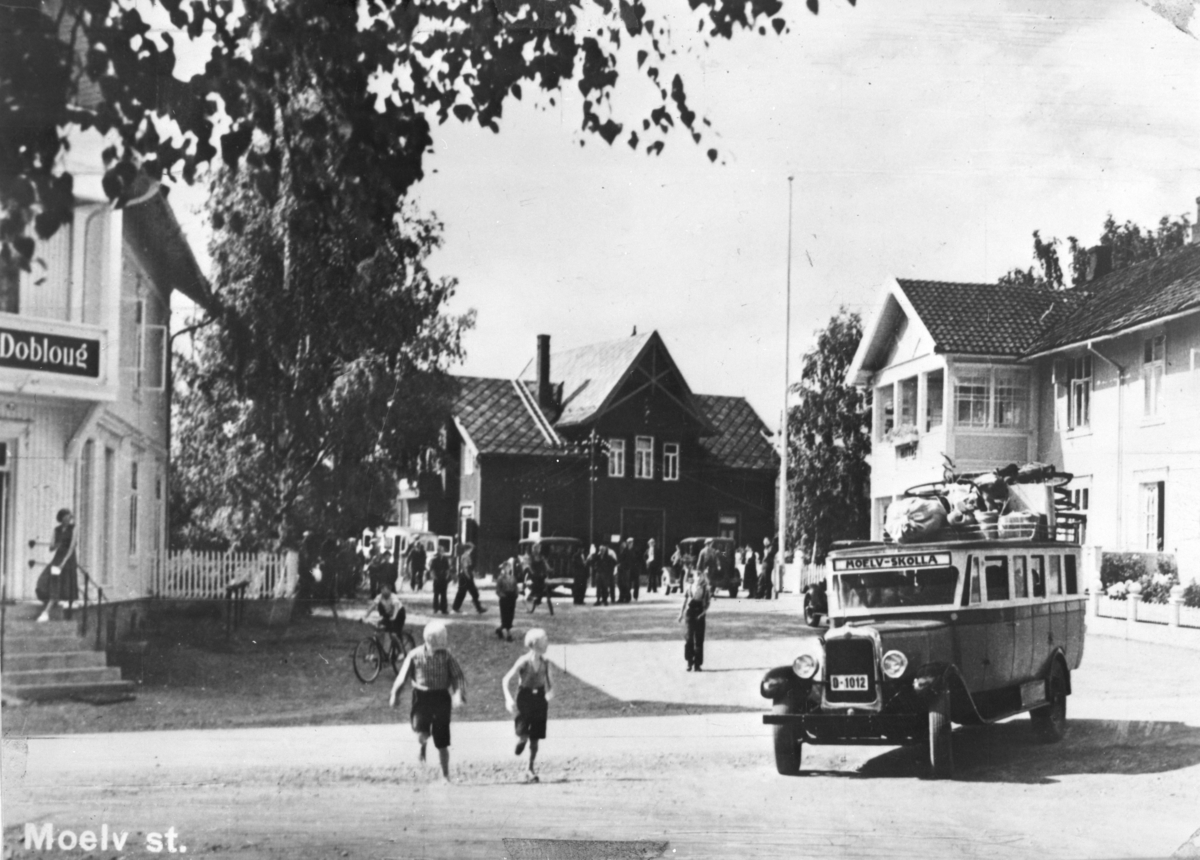 Ukjent, Moelv jernbanestasjon, Storgata, Moelv sentrum. Ca. 1935-36. Grødtliens G. M. C: buss "Moelv-Skolla", D-1012, fullastet med bagasje på veg til fjells. Guttene på bildet er Syverin Ulven og Johs. Løkkesveen. ..