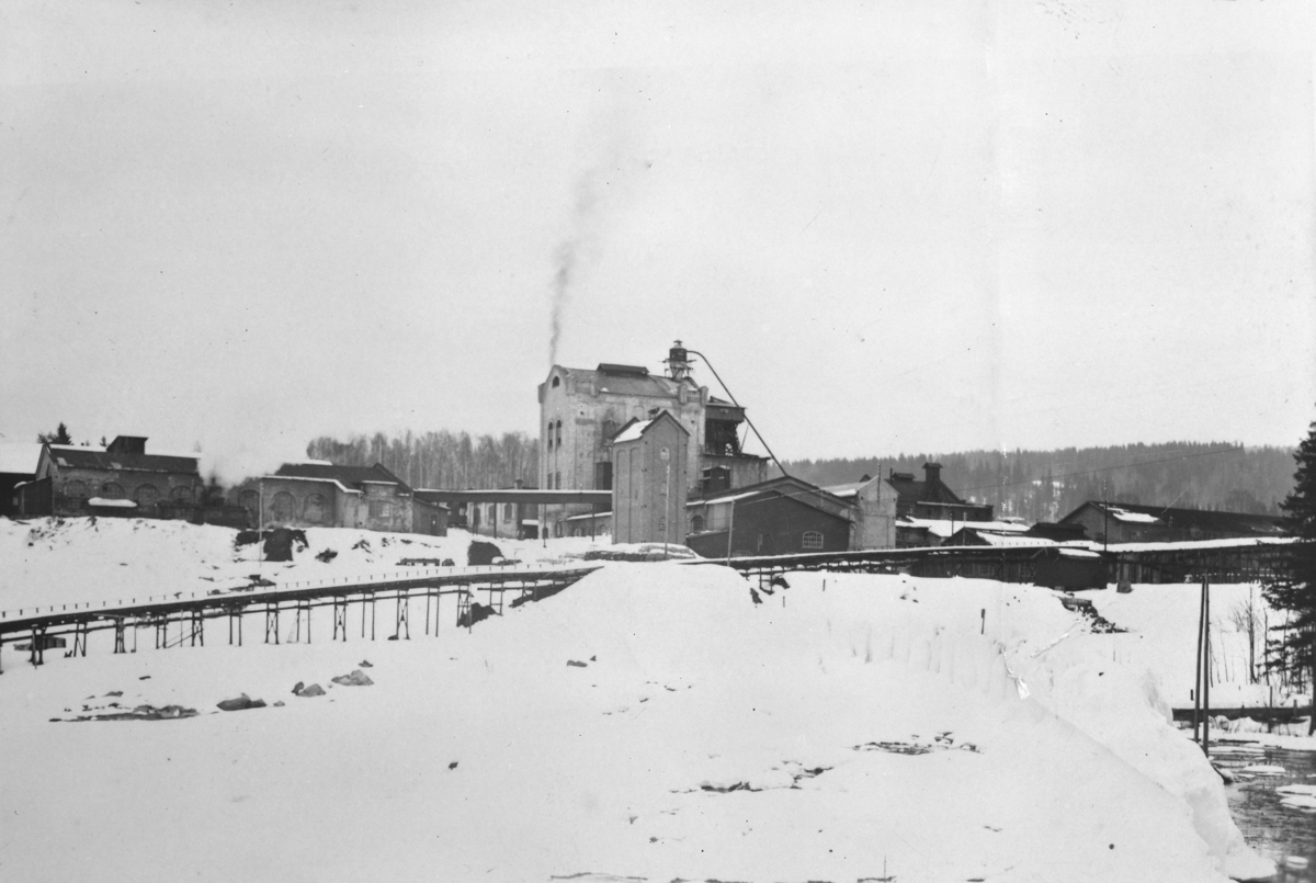 Ukjent, Moelv Cellulosefabrikk, vinter. Treforedlingsindustri. 1920-årene. Tømmeret kommer over kjerraten fra tømmerlageret i Mjøsa, for kutting, kløyving og oppmåling. Stokkene kommer inn fra venstre til renseriet. Den høge bygningen var kokeriet der "hollenderne" sto. Det lange røret opp til en syklon på…