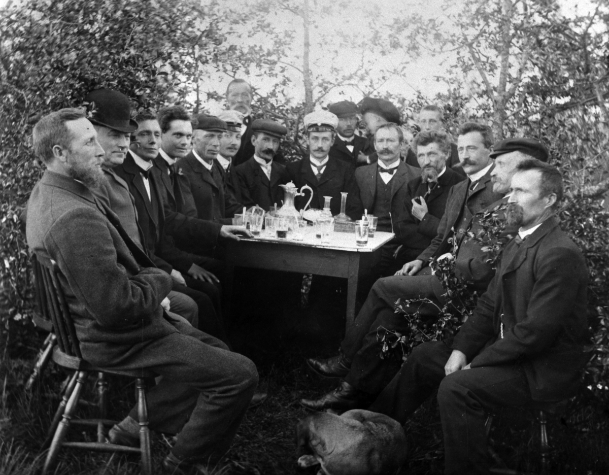 Normann Helger, TANGEN MANNSKOR SAMLET I LYSTIG LAG (PÅ HELSET ØVRE? )UTENDØRS, GLASS, KARAFLER, TANGEN, TIL HØYRE: TILLE SKOGSRUD, NR. 3 FRA HØYRE: JOHAN SKOGSRUD, 4 FRA HØYRE: SYVER FOSS, TIL VENSTRE: DIRIGENT SAKSRUD, Nr. 5 f. v. Christofer Skaven.