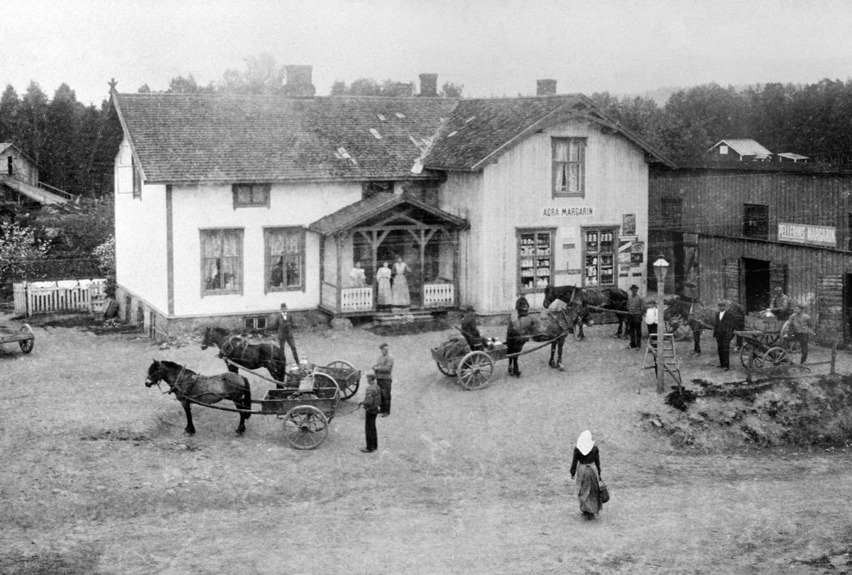 Anno Domkirkeodden, EKSTERIØR: TANGEN LANDHANDLERI, MANGE HESTER MED LASS, FOLK, TANGEN, På butikkveggen ser vi skilt for norske "Agra margarin". På lagerbygget markedføres svenske "Pellerins margarin". Karene med hesteskyss har kjørt melka til meieriet som ligger vis a vis butikken. Kona i forgrunnen skal være Berthe…