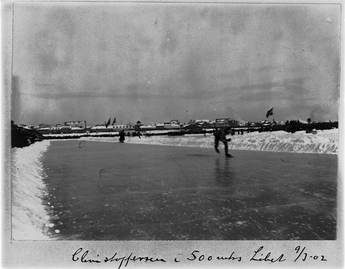 Johan Albert Steen, SKØYTELØP PÅ MJØSISEN, CHRISTOFFERSEN I 500 METERS LØPET, TRIBUNE, PUBLIKUM, FLAGG, 9-3-1902, BEBYGGELSEN I STRANDGATA, HAMAR I BAKGRUNNEN.