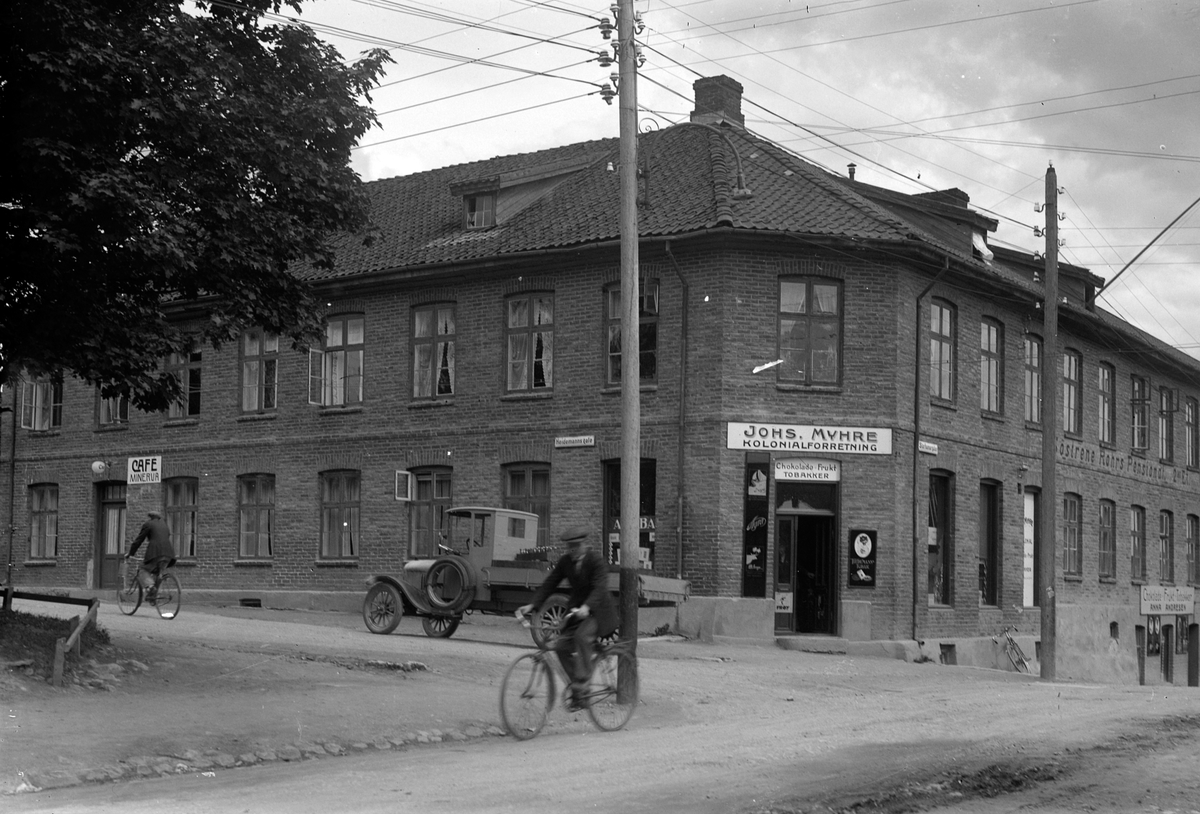 Oscar Løberg, Johs Myhre kolonialforretning, Cafe Minerva, Søstrene Røhrs Pensjonat, Storhamargata, Heidemannsgate, Hamar. Syklister, lastebil, reklameskilt. Bygningen ble satt opp av Knut Morterud da byen var ny. Søstrene Røhr drev pensjonat i annen etasje fra ca. 1890 til 1938. En periode drev de også kaféen t…