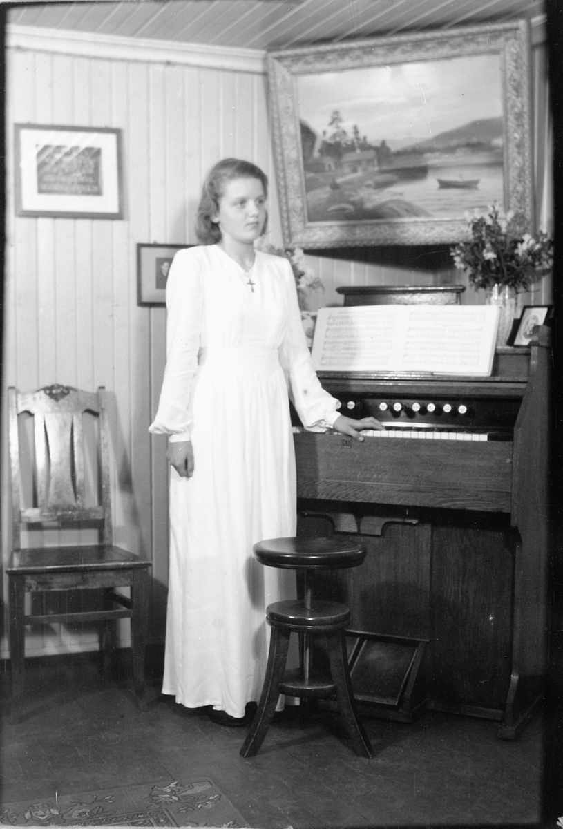 Håkon Prestkværn, Interiør, stue, konfirmasjon, Sveum. Konfirmanten som er Randi Sveum (1933-1998) står ved et orgel.