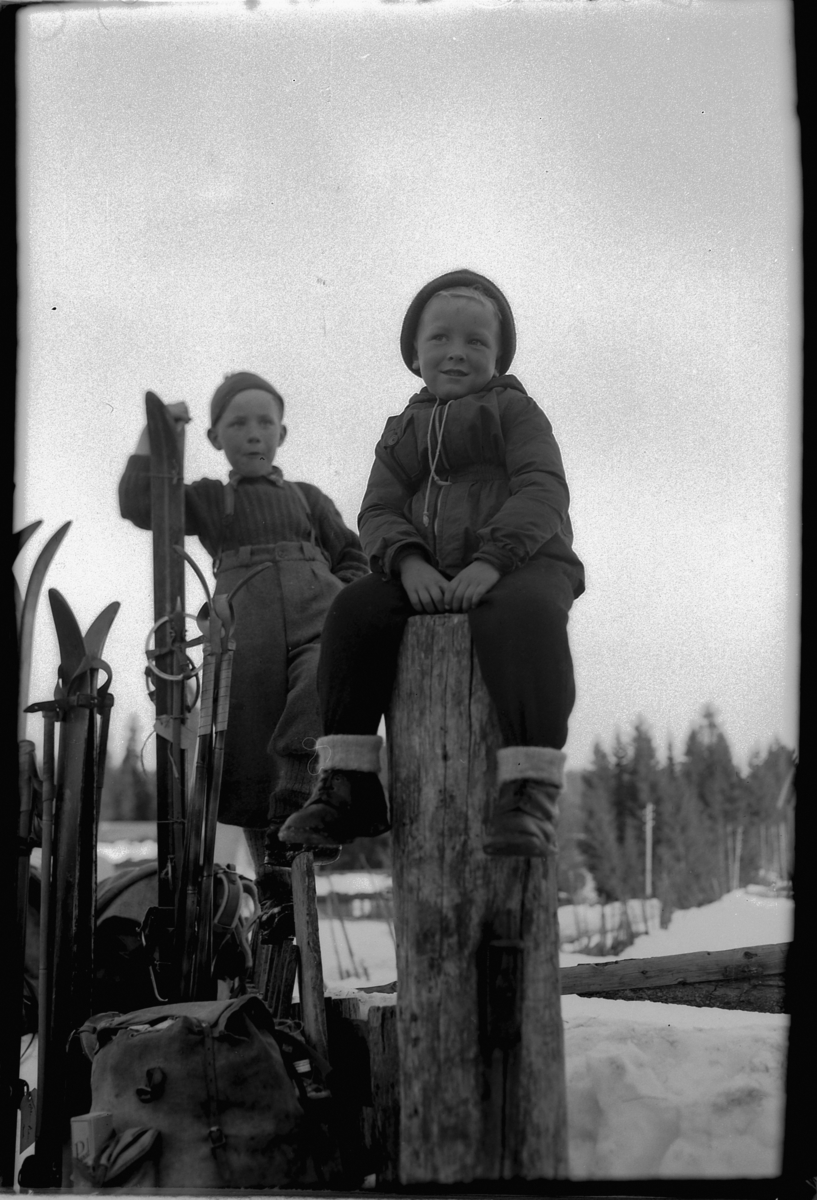 Håkon Prestkværn, To ukjente barn sitter på et gjerde med ski og sekker.