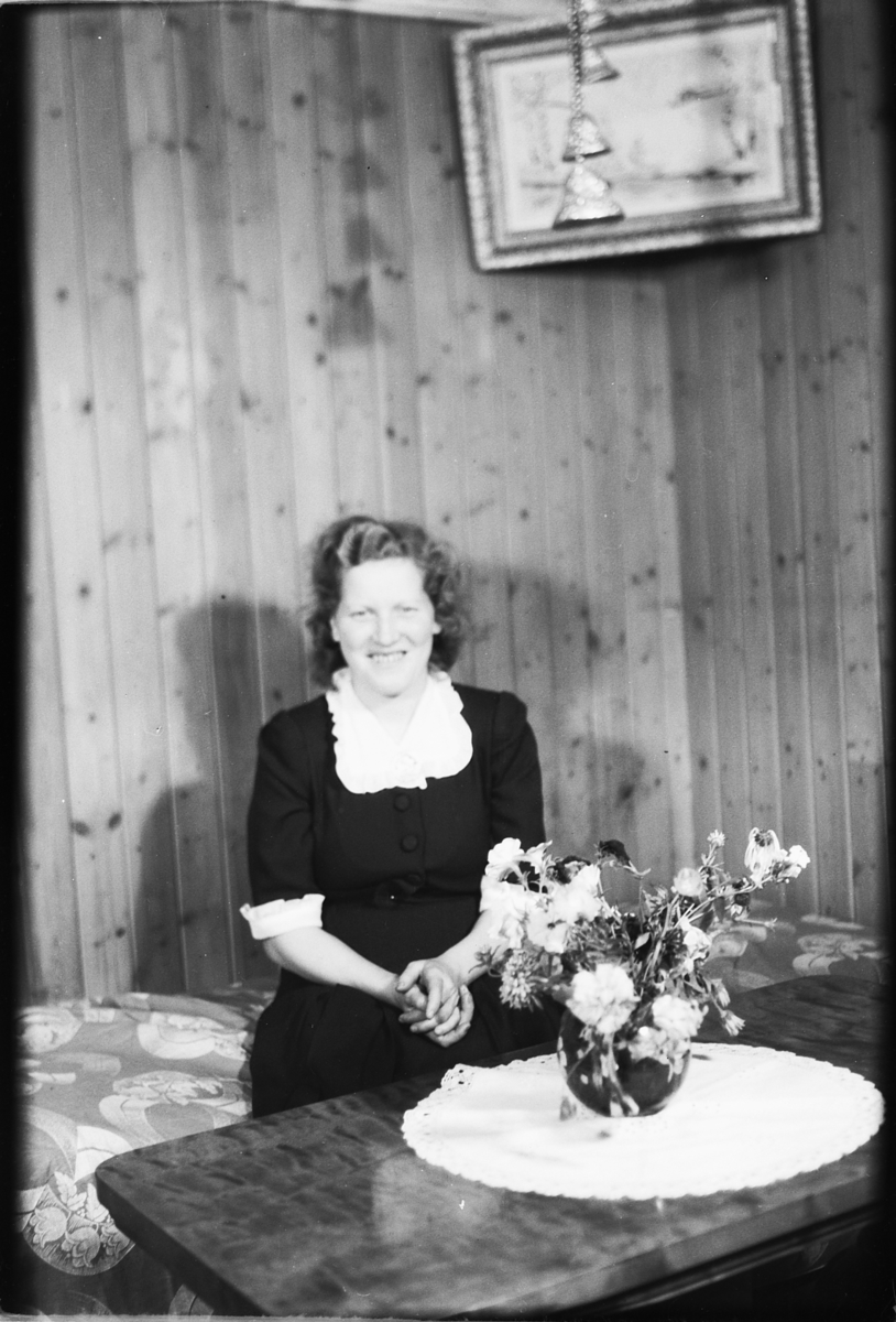 Håkon Prestkværn, Interiør, stue. Ukjent kvinne i en sofa foran bord med blomster.