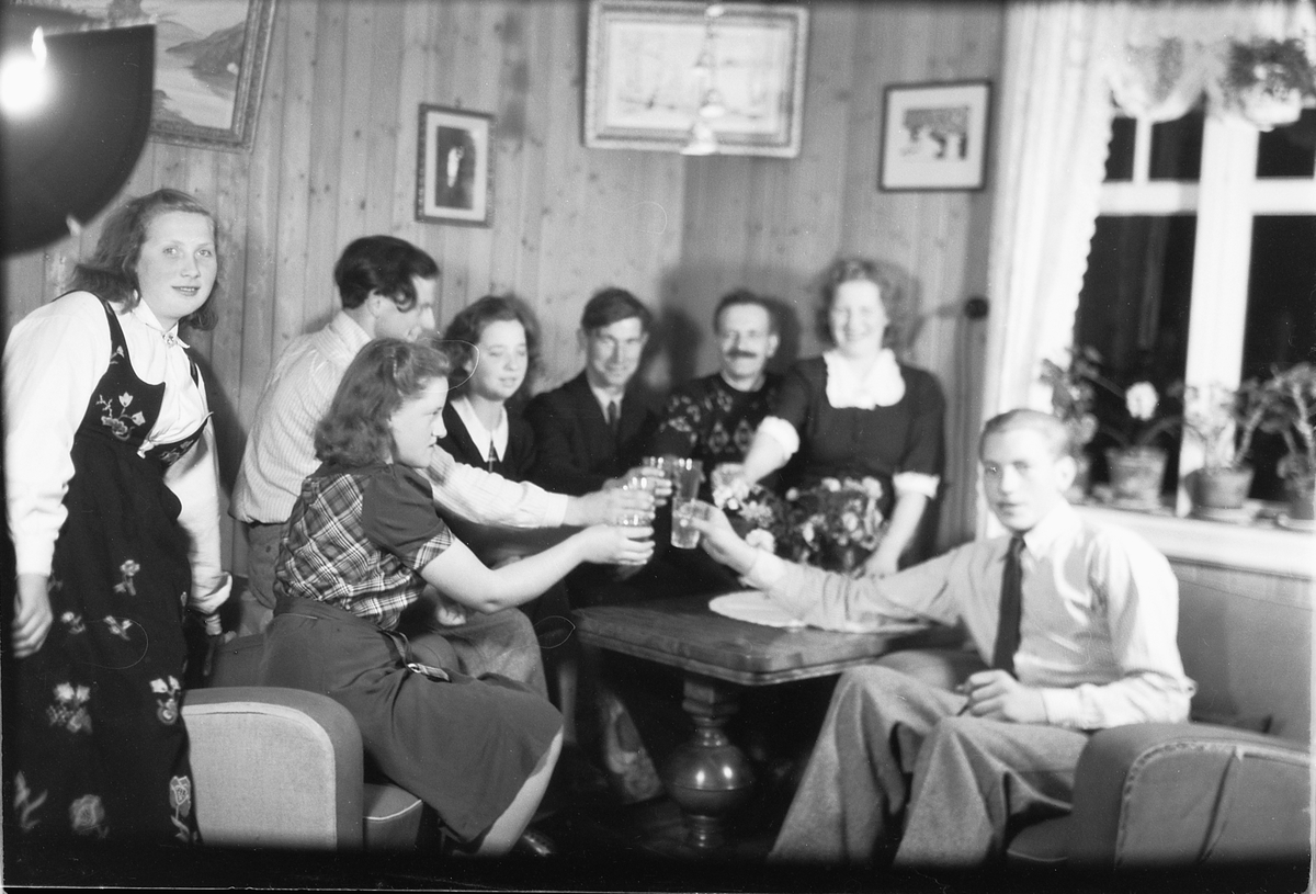 Håkon Prestkværn, Interiør, stue. Ukjent familiegruppe skåler rundt et bord. Tv en kvinne i bunad.