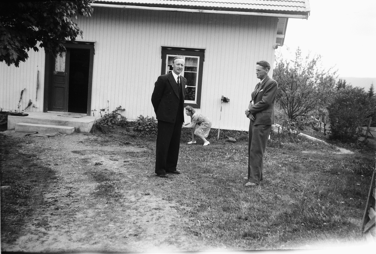 Håkon Prestkværn, To ukjente menn med en kvinne i bakgrunnen står foran et hus.