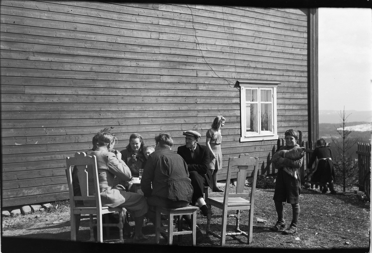 Håkon Prestkværn, Gruppe kvinner, menn og barn samlet rundt et bord i husveggen.