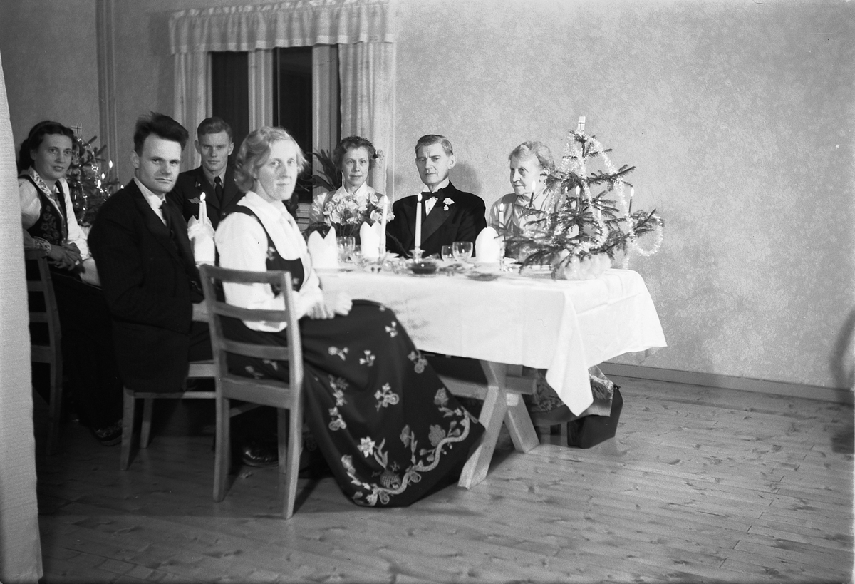Håkon Prestkværn, Interiør, stue. Ukjent gruppe mennesker rundt et bord med kvinner i bunad.