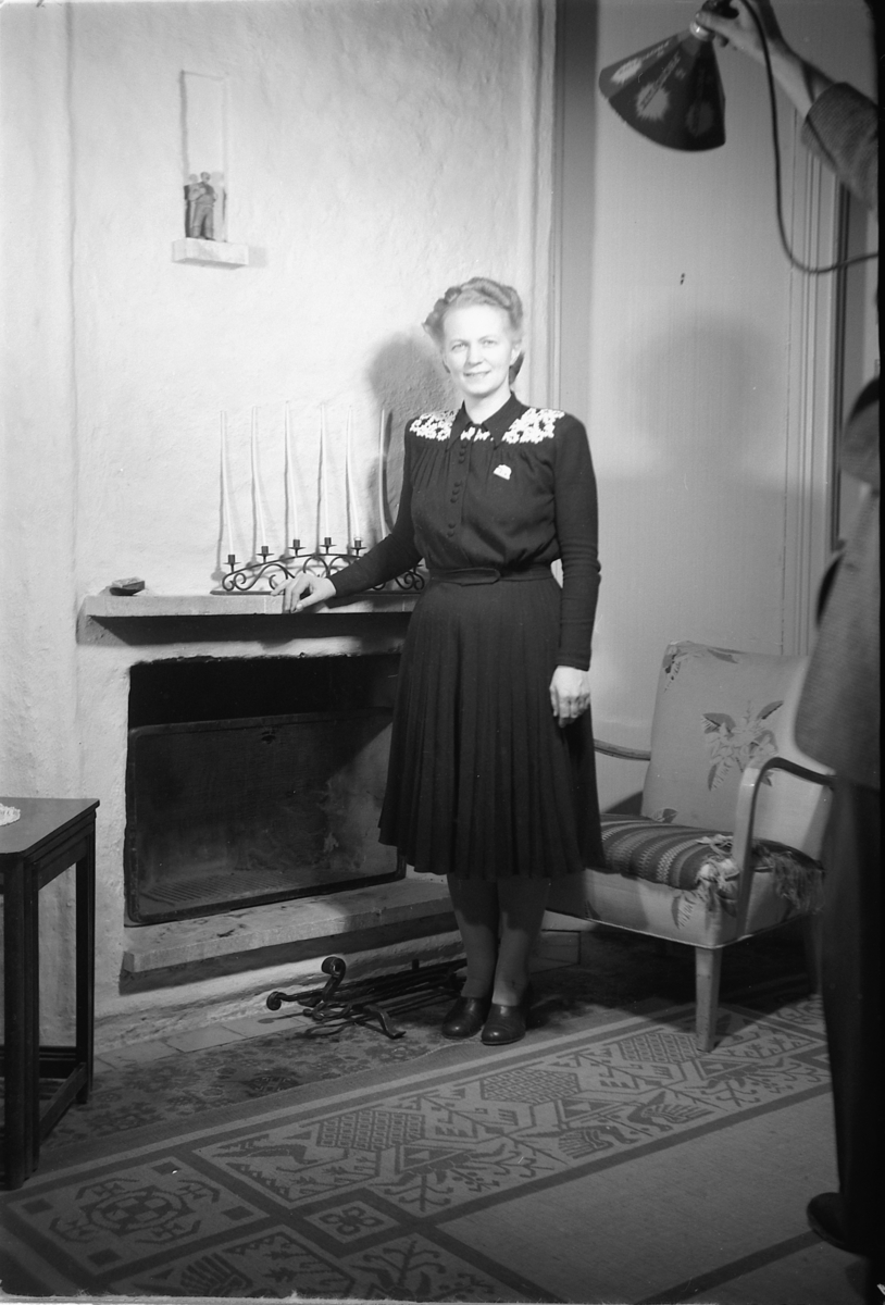 Håkon Prestkværn, Interiør, stue. Kvinne står ved en peis.