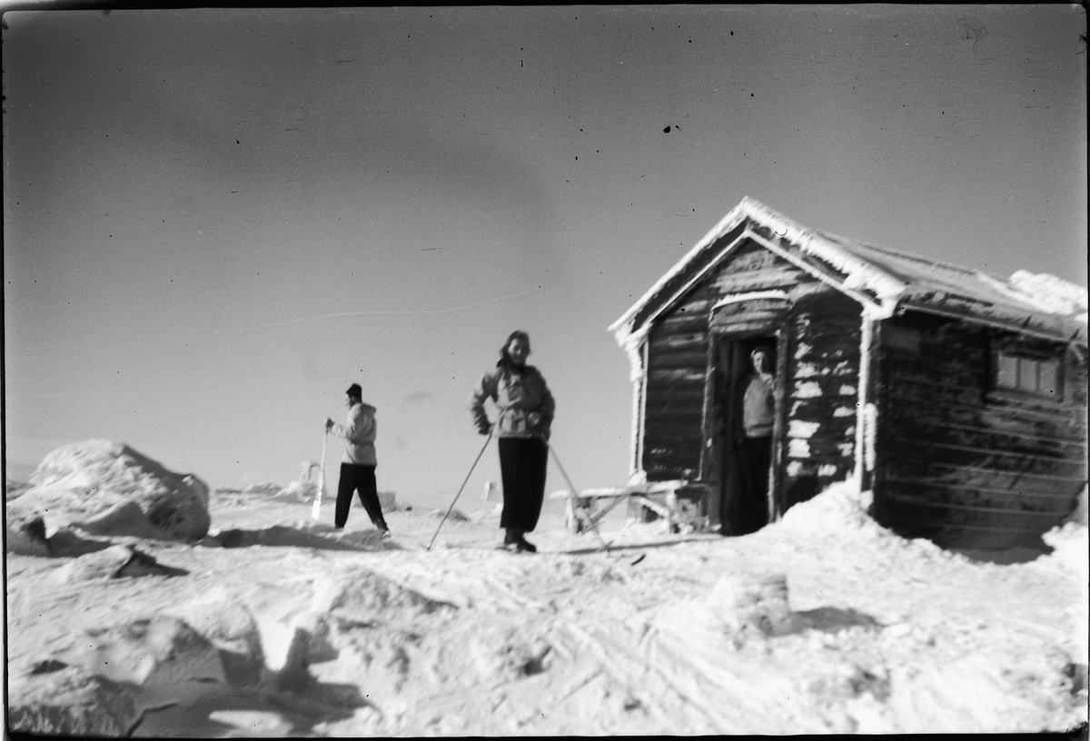 Håkon Prestkværn, To skigåere og en mann i døråpningen til ei hytte.
