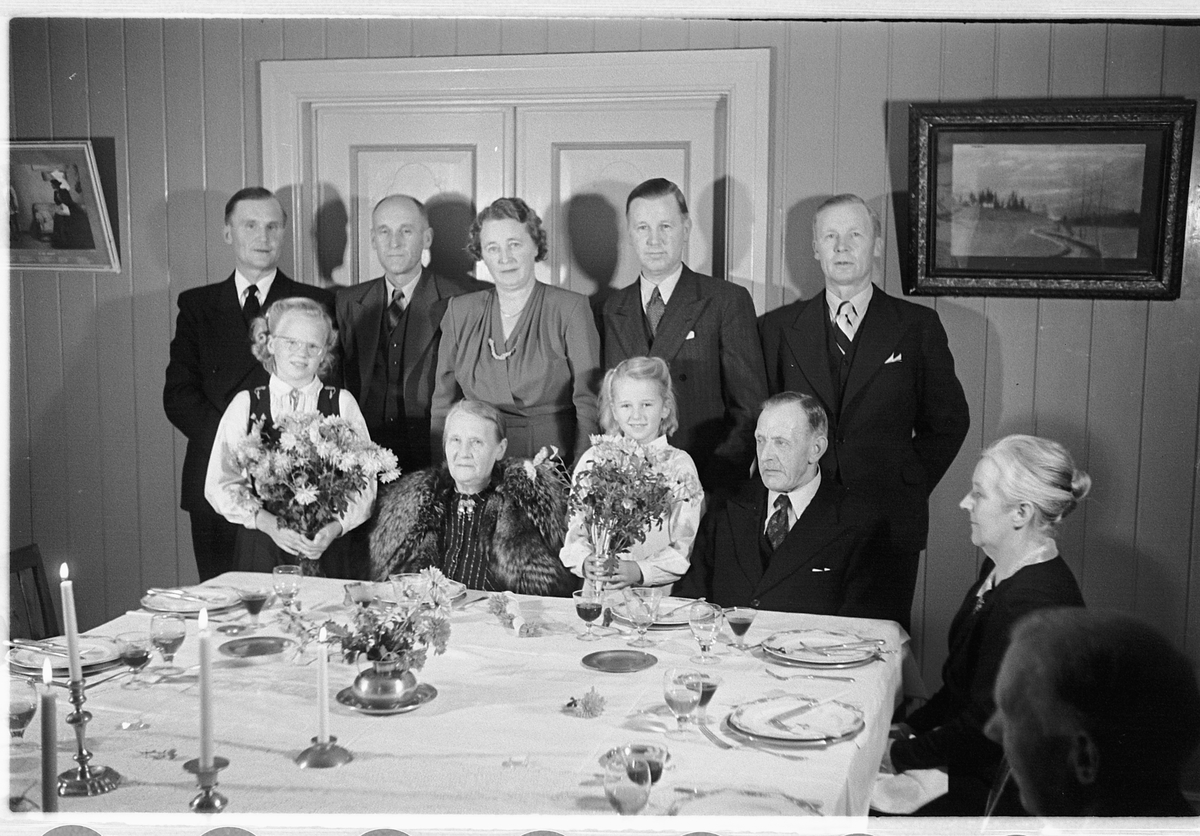 Håkon Prestkværn, Interiør, stue. Familiegruppe samlet ved et dekket bord.